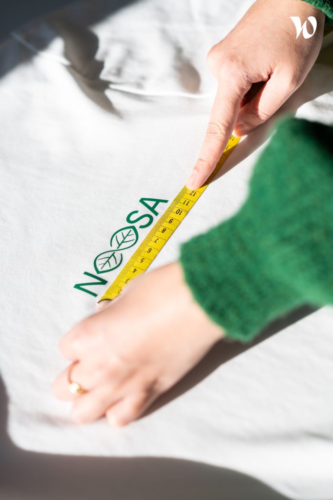 Hands measuring the size of the NOOSA™ logo on a white t-shirt, end-products of NOOSA™.