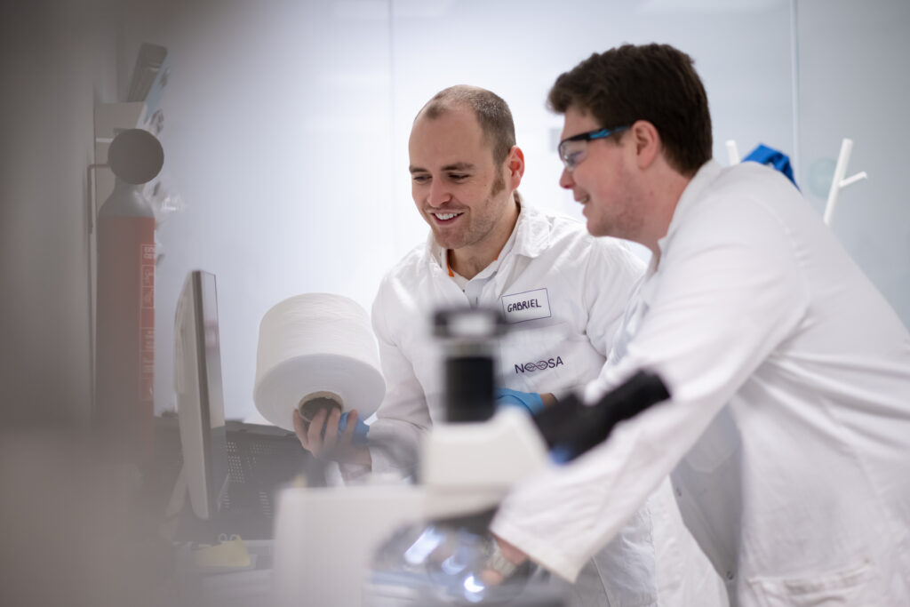 Two NOOSA™ scientists wearing white lab coats and safety gear are engaged in a discussion. One of them, identified by a name tag as "Gabriel," is holding a large roll of material. They are standing in a laboratory with equipment and instruments in the background.