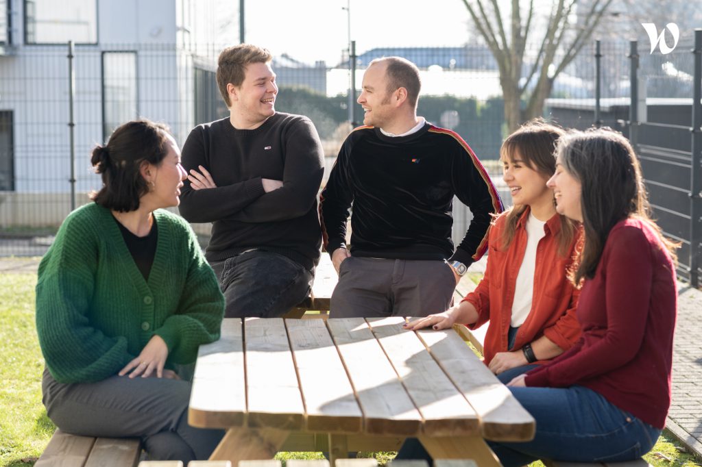 Five NOOSA™ team members sitting and standing around a wooden picnic table outdoors, smiling and talking to each other