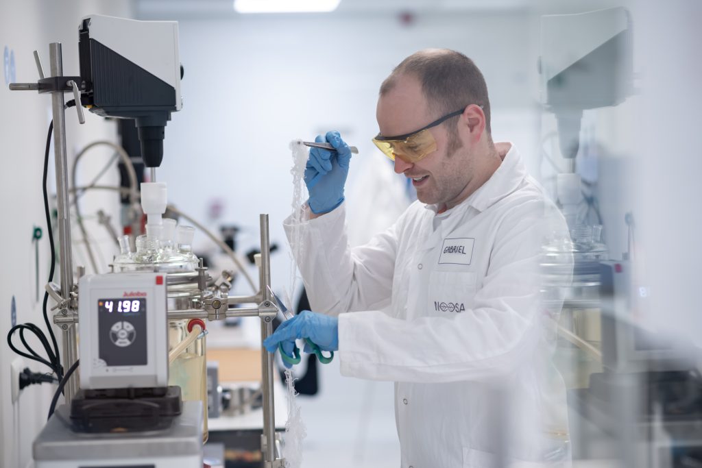 Gabriel, NOOSA™ Phd researcher in a lab coat and safety glasses works with laboratory equipment, holding a piece of fiber with tweezers