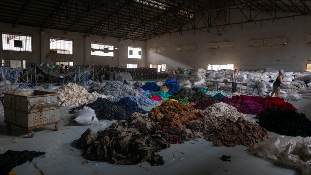 Interior of a large warehouse filled with piles of various colored fabrics spread out on the floor. Two people are seen working among the fabric piles, with large bags of textiles stacked against the walls and some industrial equipment visible.