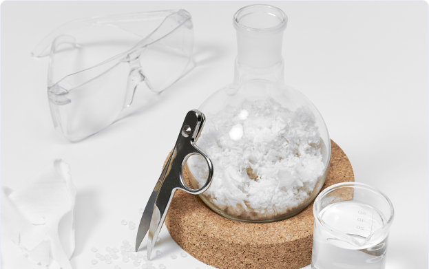 A laboratory setup featuring clear safety goggles, a glass beaker filled with NOOSA™ fiber a small glass container with clear liquid, and a pair of scissors. The items are placed on a cork mat, and there are some small white granules and a piece of torn white fabric nearby.