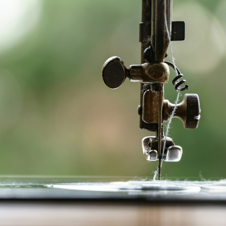 A sewing machine needle with a thread, focused on the mechanism. The background is blurred with soft green and beige hues.