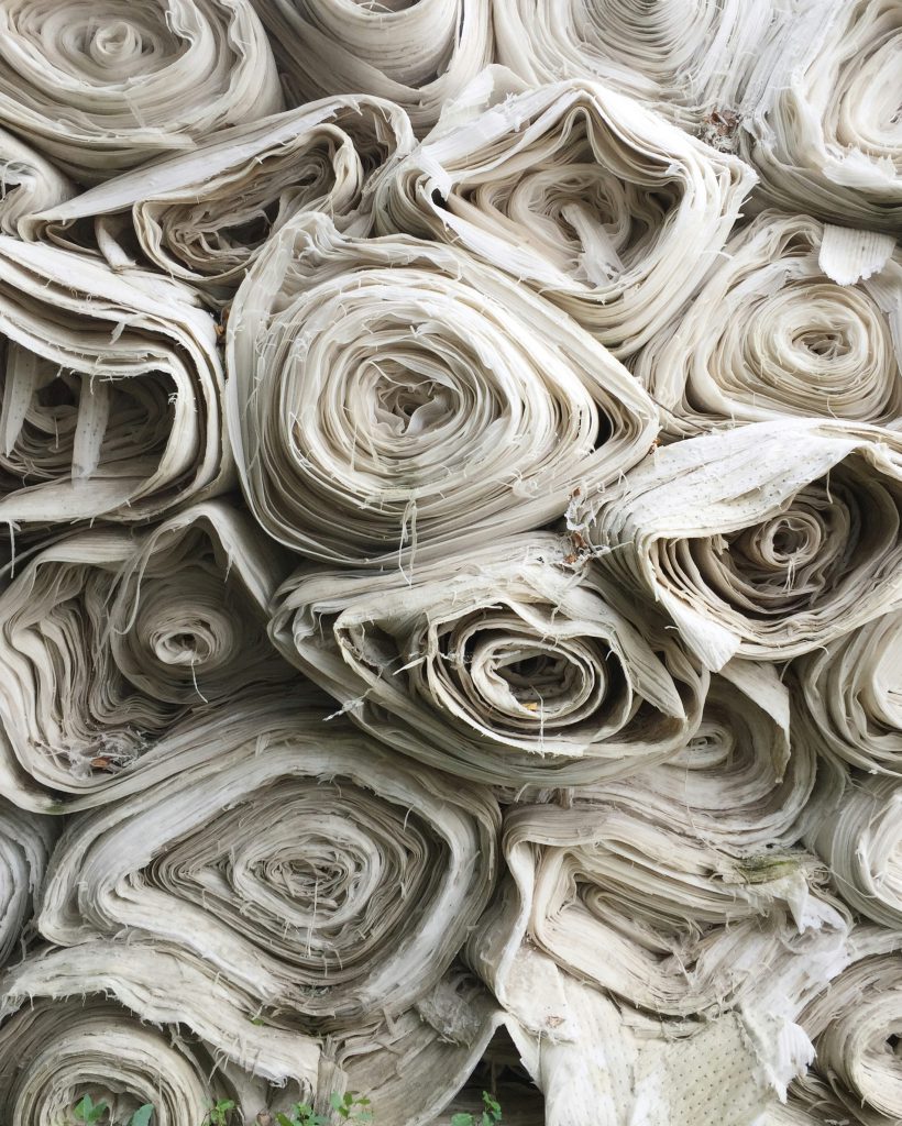 A large pile of rolled white fabric, with each roll tightly wound and stacked on top of one another. The edges of the fabric are slightly frayed.