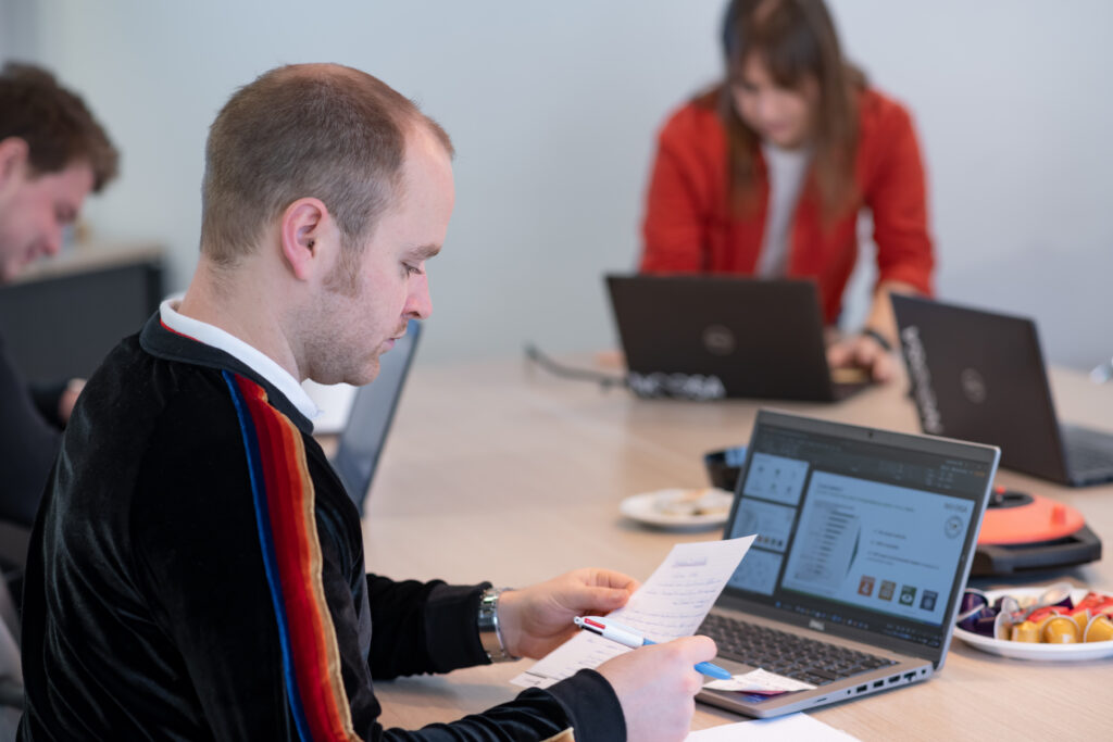 Gabriel, a NOOSA™ team member is seated at a desk, focusing on a sheet of paper while working on a laptop. In the background, two other people are also working on laptops. The desk has a bowl of snacks and other office items.